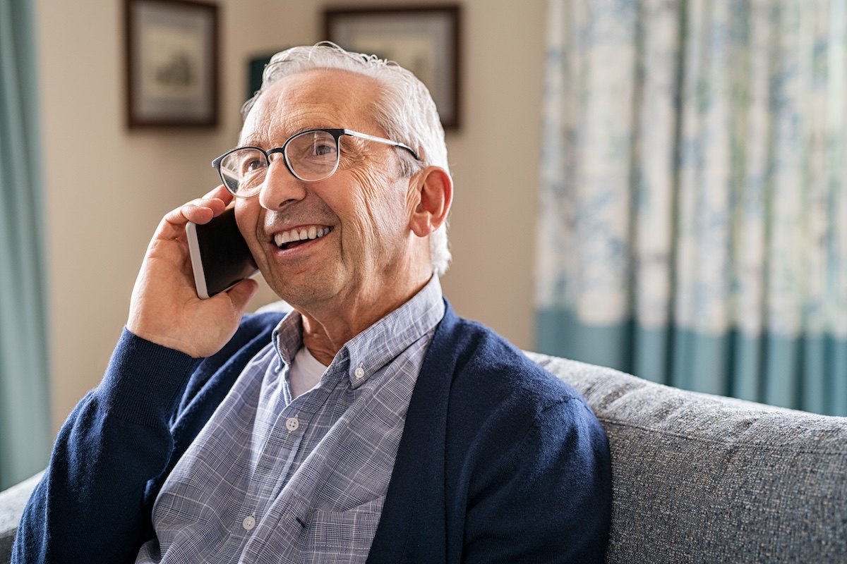 Smiling old man talking over smartphone while relaxing at home. Senior man after retirement using smart phone to connect with friends and family. Carefree elder talking over mobile phone with her daughter while sitting on couch and looking up.
